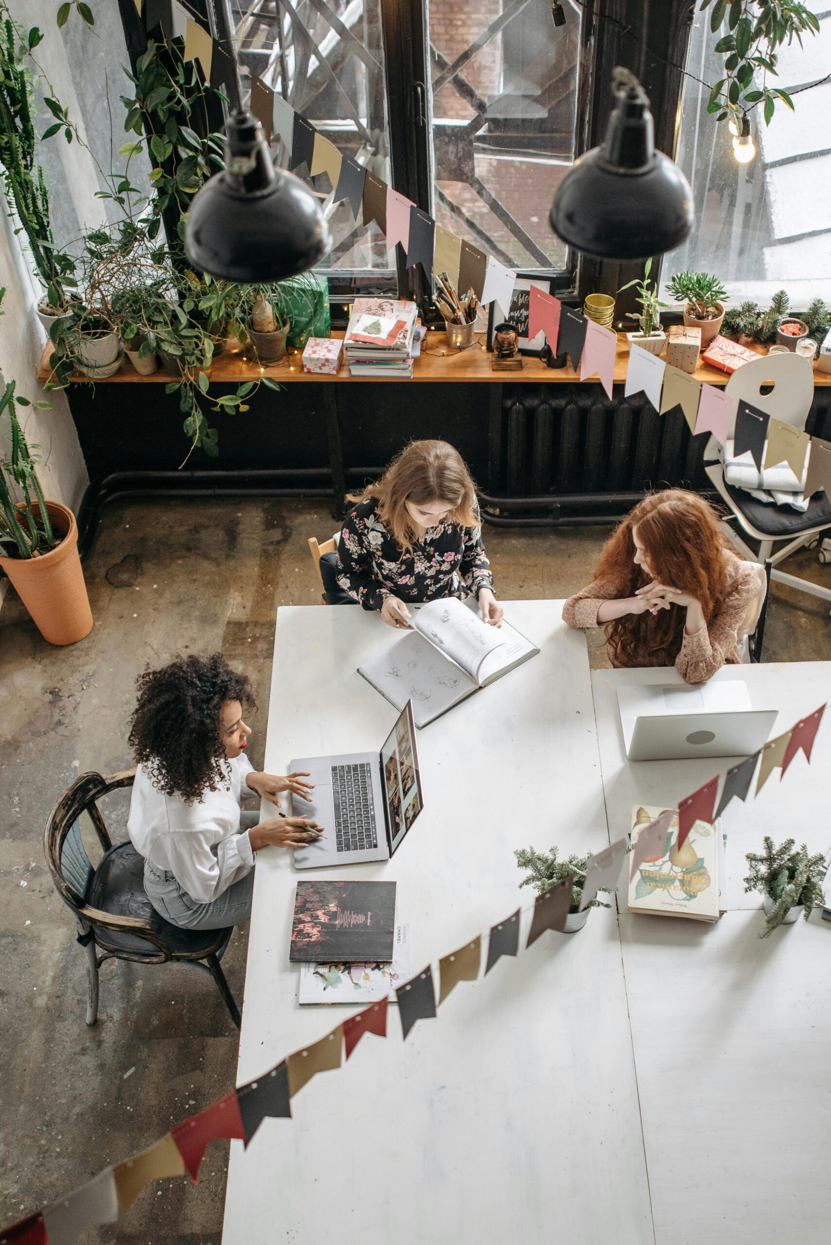 Three women collaborate creatively in a cozy indoor workspace filled with plants.