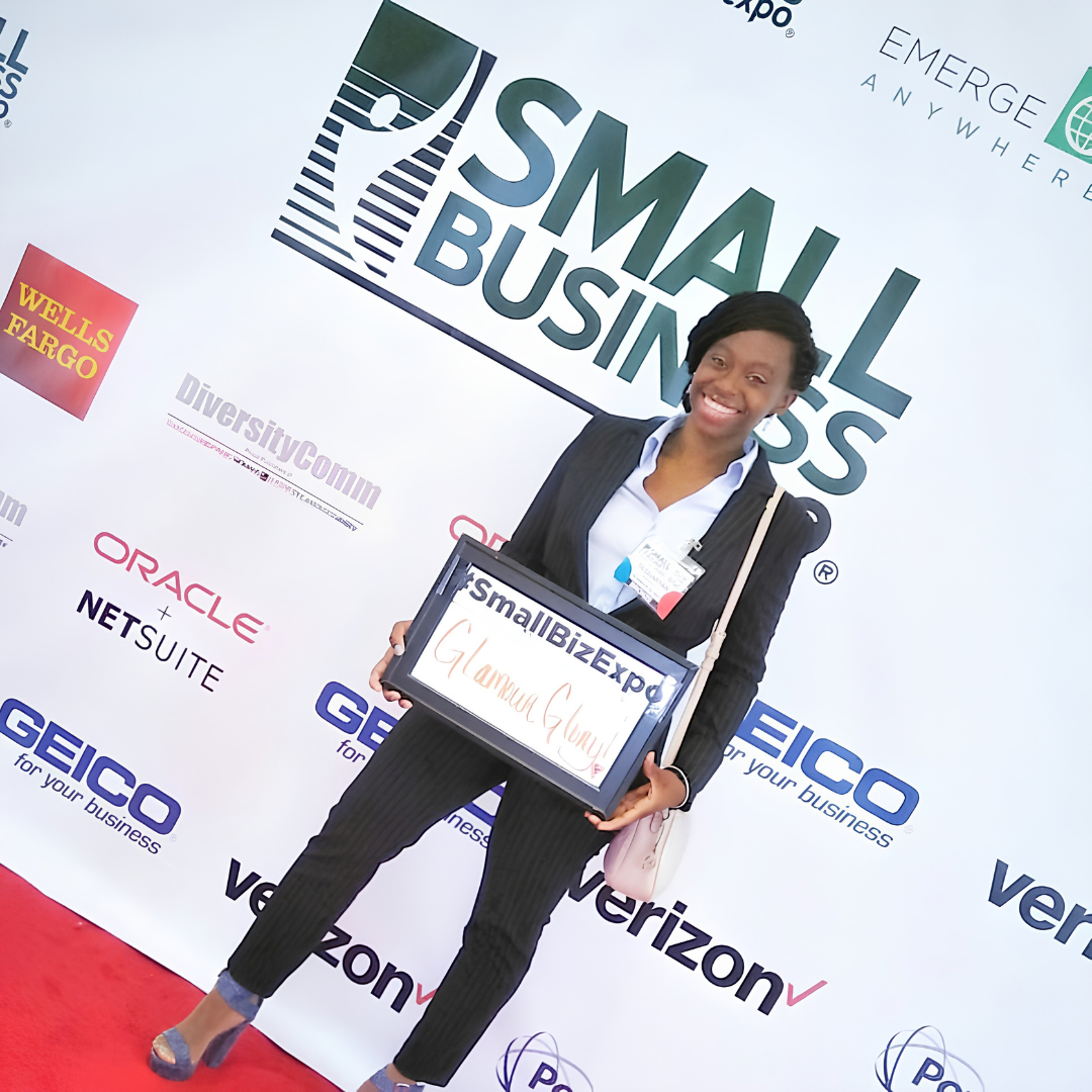 Ta'Quaryan in a black dress holding a framed Glamour Glory sign at her Small Business Expo booth